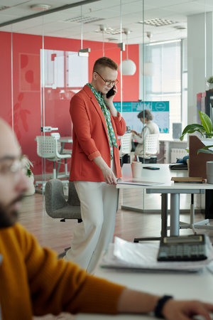Professional businesswoman engaging in conversation on smartphone while standing in modern office,...