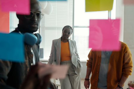 Three colleagues brainstorming during meeting, focusing on colorful sticky notes on glass wall,...