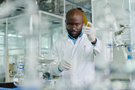 Young male clinician in eyeglasses, lab coat and protective gloves dropping liquid substance into flask during chemical experimentの写真素材