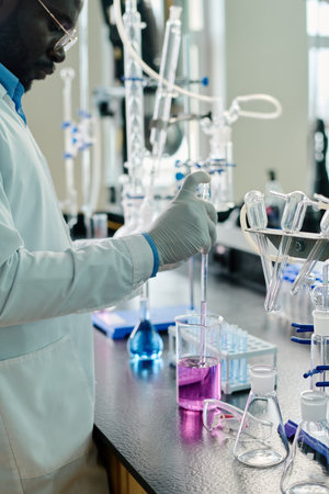 Cropped shot of gloved male scientist in lab coat standing by workplace with glassware containing liquid substances during clinical trialの写真素材