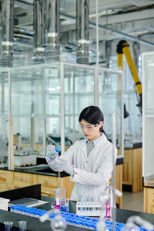 Chinese female researcher in lab coat, protective eyeglasses and gloves carrying out chemical...