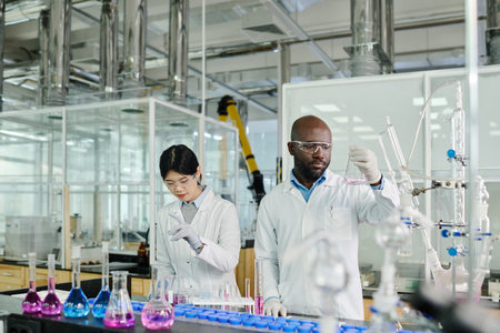 Female clinician mixing two substances in glassware while her male colleague studying its chemical...