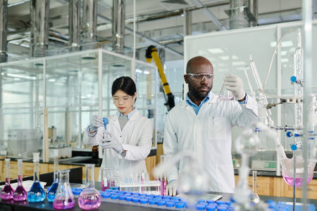 Young serious male scientist in lab coat looking at liquid in beaker while standing next to female...