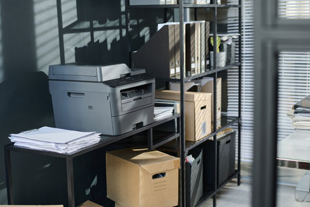 Plastic copier machine standing on desk with stack of paper documents by stand with shelves with cardboard boxes and folders with billsの写真素材