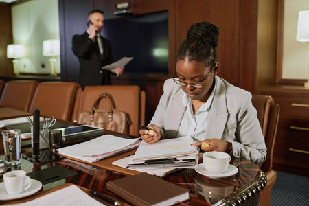 Serious young female solicitor bending over folder and paper documents while sitting by table in...