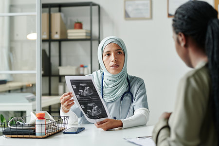 Young confident female gynecologist in hijab showing her pregnant patient sonogram of baby while...