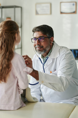 Smiling mature male pediatrician with stethoscope sitting in front of little patient and examining...