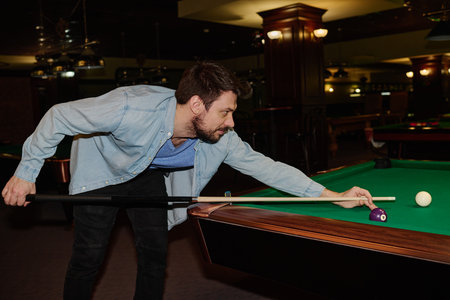 Side view of young man in black jeans and denim shirt standing by wide pool table while hitting one...