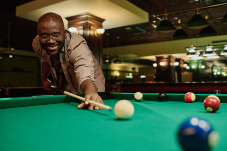 Young smiling man in casualwear and eyeglasses hitting white billiard ball with cue during game...