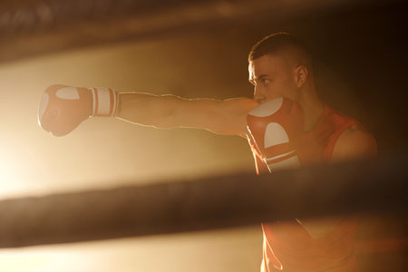 Young boxer holding left arm bent in elbow by his face while kicking rival with right hand in boxing glove during training on ringの写真素材