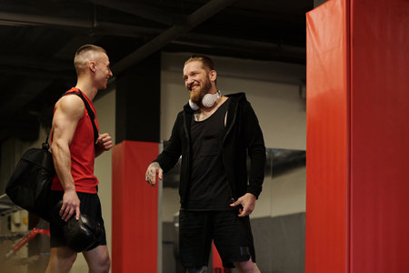 Two young cheerful male athletes in sportswear looking at one another during conversation while standing in gym before trainingの写真素材