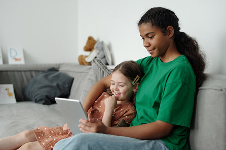 Cute teenage girl in casualwear sitting on couch next to adorable child and embracing her while both...