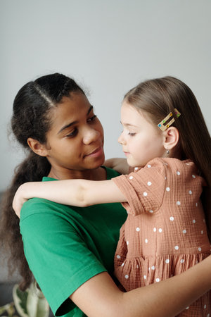 Pretty teenage girl in green t-shirt looking at adorable little child on her hands while spending time with her in home environmentの写真素材