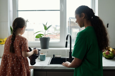 Rear view of cute little girl in casual dress standing by kitchen counter and drying clean dishes with soft towel while helping baby sitterの写真素材