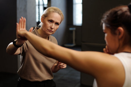 Mature boxing trainer looking at young female learner keeping arm with punch stretched during kick while practicing exercises in gymの写真素材