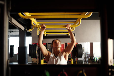 Young active woman in sportswear looking upwards while holding by yellow metallic bar of sports...