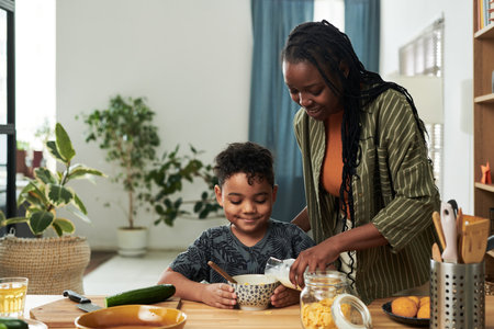 Happy young African American woman pouring fresh milk from bottle in bowl with cornflakes while making breakfast for her little sonの写真素材