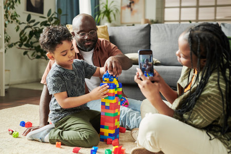 African American child and man sitting in front of young woman with mobile phone taking video of her family playing constructorの写真素材