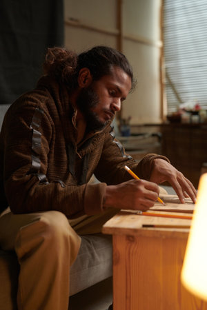 Man concentrating on drawing while sitting at wooden table in cozy art studio with soft light...