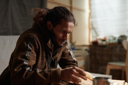 Man sitting in workshop deep in thought with tools spread on table around him creating atmosphere of focus and creativityの写真素材