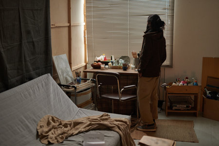 Woman standing in cozy bedroom looking out window with thoughtful expression, bedroom filled with...