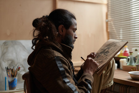 Artist with long hair and beard working meticulously on sketch while seated in studio filled with art supplies, brushes, and sketches around sketchbookの写真素材