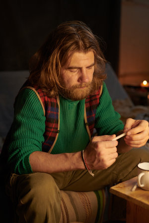 Man with long hair and beard concentrating while inspecting small object in his hands, sitting on patterned couch in cozy indoor setting with dim lightingの写真素材