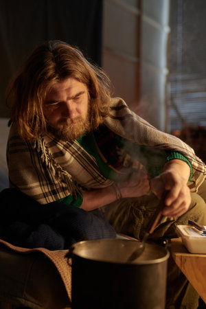 Bearded man with long hair stirring pot over heat in rustic indoor setting with daylight pouring in...