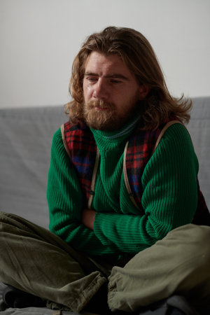 Man with shoulder-length hair and beard sitting cross-legged on a couch with arms crossed wearing...
