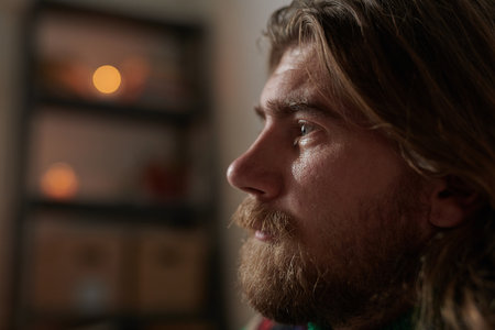 Side profile of bearded man with long hair contemplating, soft indoor glow in background, creating an intimate and thoughtful atmosphereの写真素材