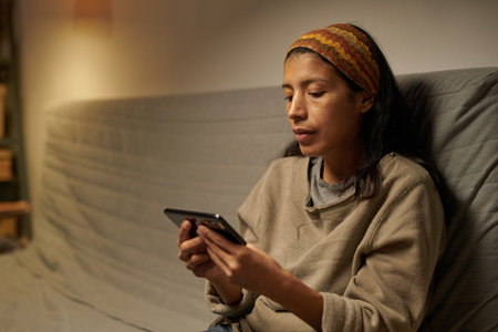 Hispanic woman sitting on sofa using smartphone while surrounded by dimly lit cozy room, wearing casual clothes and focused on screenの写真素材
