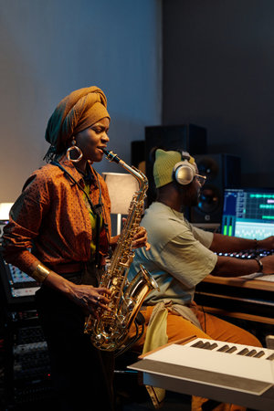 Jazz musician in recording studio, playing saxophone while producer adjusts controls on mixing board...