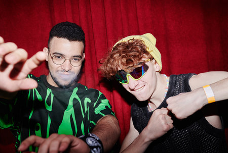 Young intercultural men in funky apparel standing and posing in front of camera against deep red curtains in night clubの写真素材
