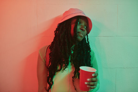 Pretty African American girl with long dreadlocks holding red cup of soda while standing by wall lit with neon light in night clubの写真素材