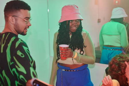 Happy African American woman in stylish attire having soda and looking at one of her friends while standing next to her boyfriendの写真素材