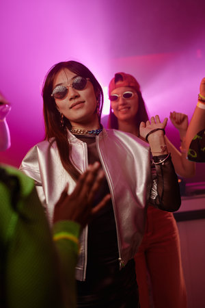 Young pretty woman in stylish sunglasses and futuristic silver jacket dancing in night club while...