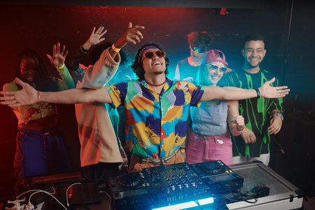 Group of young excited people in trendy attire keeping their arms raised while dancing behind happy male deejay standing by soundboardの写真素材