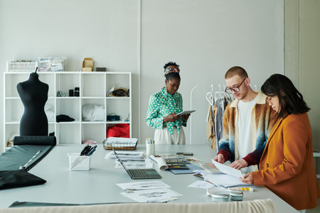 Three young intercultural coworkers standing in spacious workshop and looking though online and...