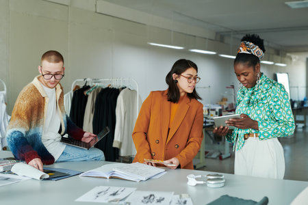 Young brunette female fashion designer looking at screen of tablet held by African American woman during presentation of new itemsの写真素材