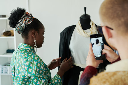 Hands of young male fashion designer with smartphone taking picture of female colleague creating new...