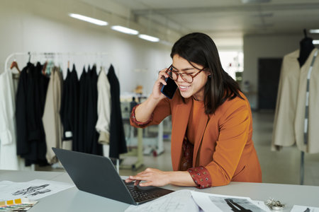 Young smiling female owner of modern atelier using laptop and consulting clients on mobile phone while bending over workplaceの写真素材