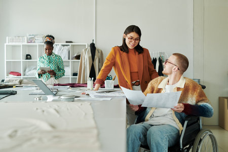 Happy young female fashion designer looking at male colleague with disability during discussion of...