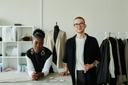Two young intercultural fashion designers in contrast casualwear looking at camera while standing by workplace with unrolled grey textileの写真素材