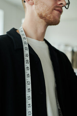 Close-up of young bearded tailor or fashion designer with measuring tape on neck and chest standing in front of camera in workshopの写真素材