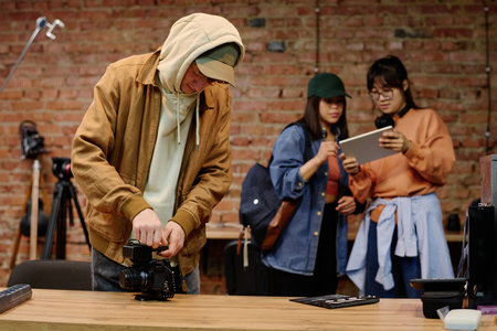 Man adjusting camera on desk while two team members in background are discussing project using tablet in modern photography studio with brick wall interiorの写真素材
