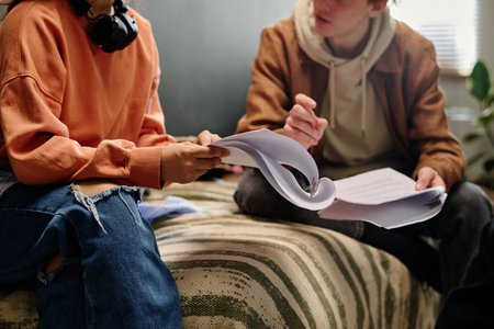 Two individuals sitting on couch and discussing notes while studying. Casual setting with focus on interaction and collaboration without direct eye contact with cameraの写真素材