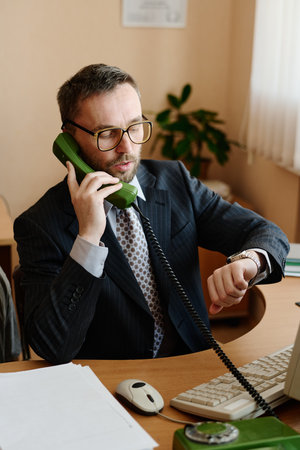 Portrait of a Caucasian businessman wearing glasses and a suit talking on an office telephone, while sitting at a desk, looking composed and focusedの写真素材