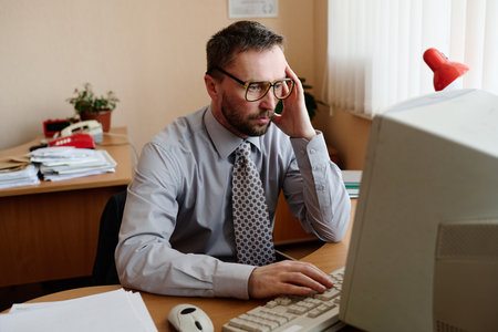 Portrait of a Caucasian man sitting at desk, using a computer in an office environment, wearing...
