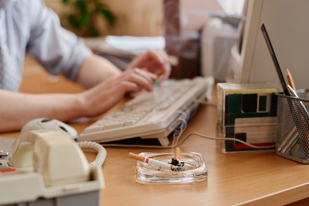 Close-up of hands typing on keyboard with ashtray containing cigarette on desk. Office supplies and...
