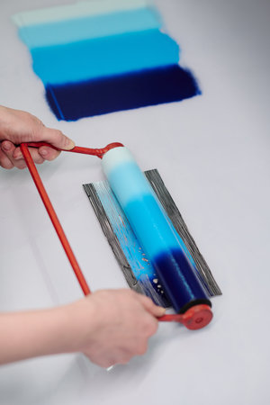 High angle of female artisan hands holding by red handle of roller with gradient blue paint while printing new sample by workplaceの写真素材